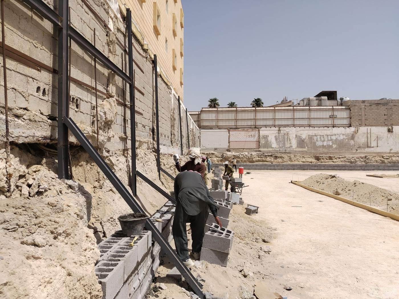 Workers laying concrete block to build a wall beside a building on a sandy site